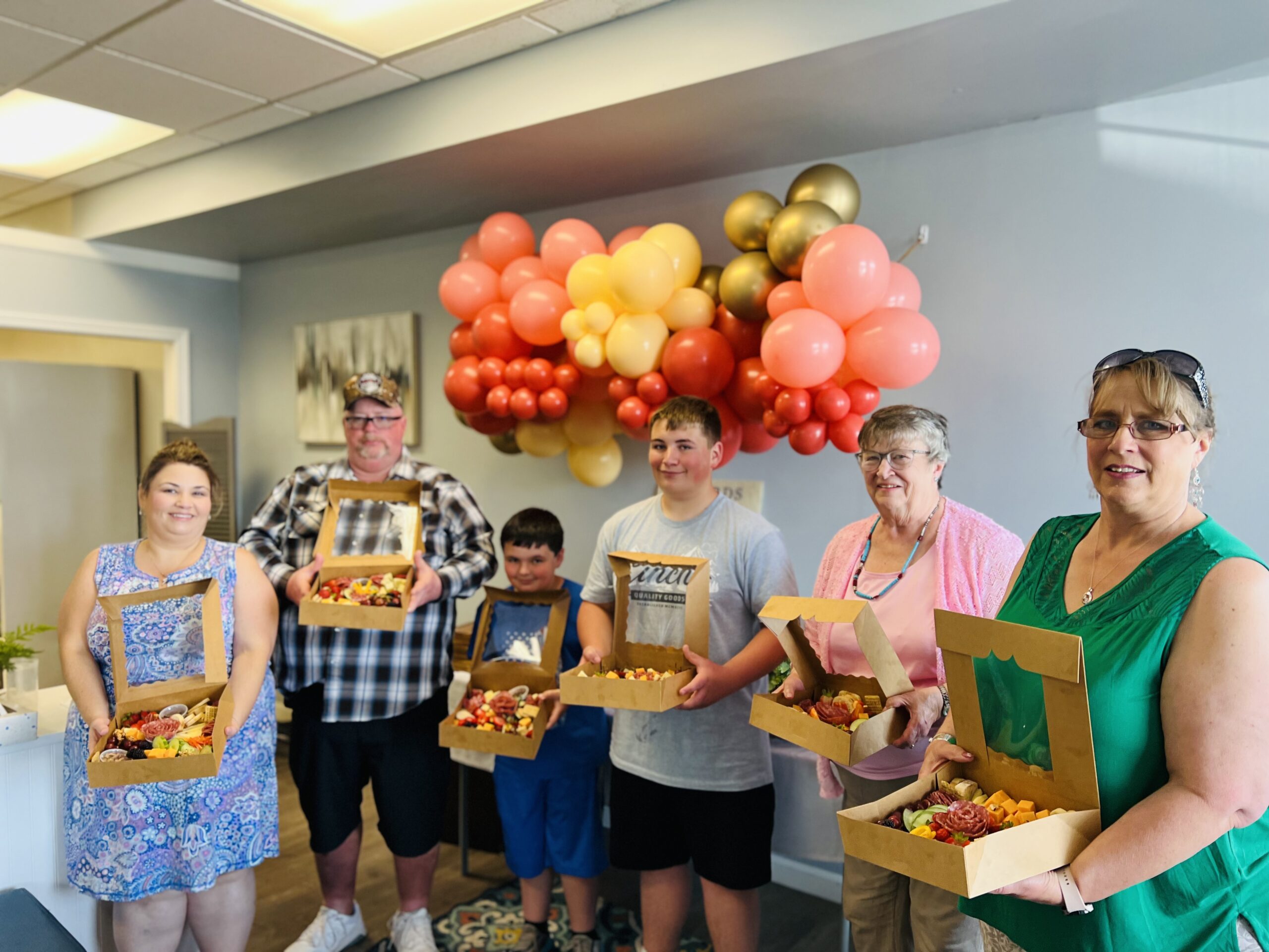 Group of smiling people holding pizza boxes under colorful balloon decorations.